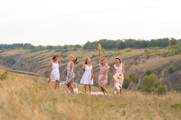 La compañía de alegres amigas se divierten juntas en un picnic en un lugar pintoresco con vista a las verdes colinas. Chicas vestidas de blanco bailando en el campo