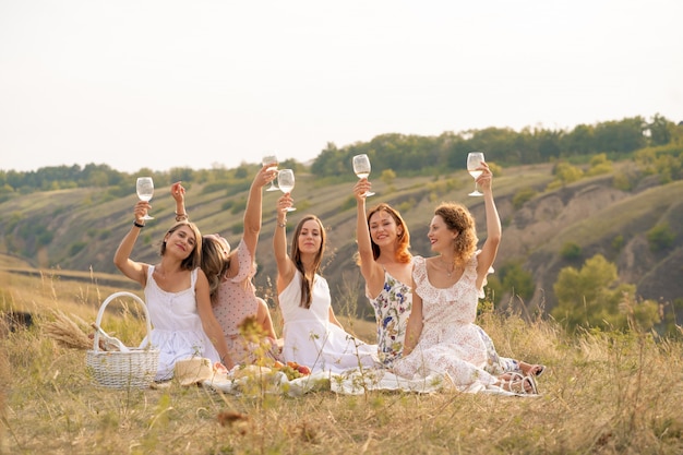 La compañía de amigas se divierte y disfruta de un picnic de verano en los jardines verdes y levanta copas con vino. Concepto de personas