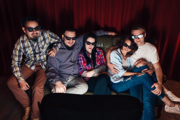 Compañía de cinco jóvenes felices en gafas 3d ven la televisión en casa