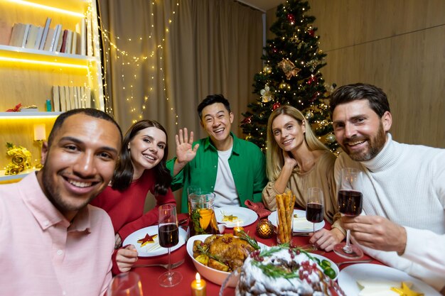 Una compañía de jóvenes interraciales sentados en una mesa festiva de año nuevo y tomándose un selfie