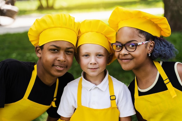 Compañía multirracial de niños cocineros con uniformes amarillos sonríen al aire libre Adolescente africano y niña negra se divierten con un niño caucásico y cocinan retrato de niños