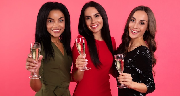 Una compañía de tres niñas brinda con copas de champán, celebrando la víspera de Año Nuevo con hermosos vestidos de color verde, negro y rojo.