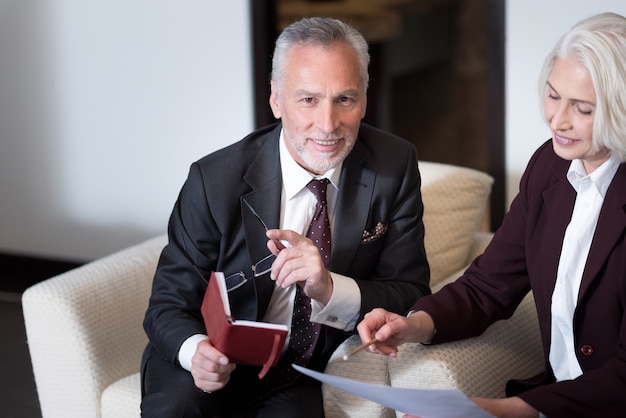 Comparta una atmósfera positiva. Encantado de hombre de negocios de edad feliz sonriendo y sentado en la oficina cerca de la empresaria mientras trabaja con su colega e intercambia ideas