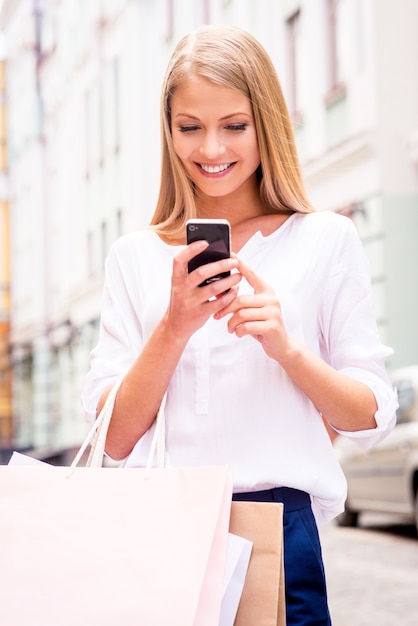 Compartiendo buenas noticias con un amigo. Primer plano de la hermosa joven sonriente sosteniendo bolsas de la compra y teléfono móvil mientras está de pie al aire libre