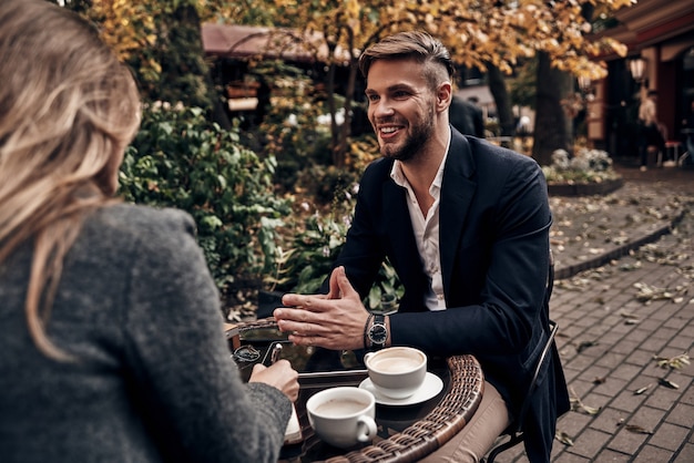 Compartiendo grandes ideas. Hombre joven guapo en ropa casual elegante sonriendo mientras tiene una conversación con una mujer joven en el restaurante al aire libre