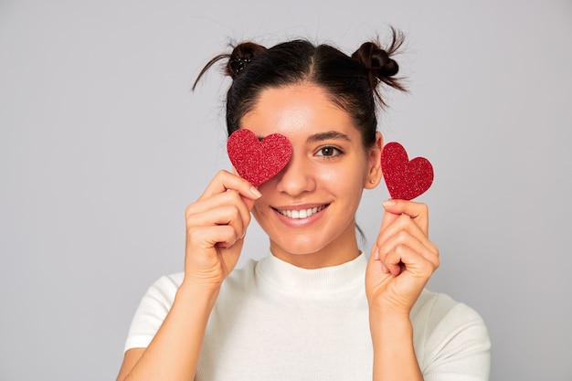 Compartilhando amor e namorados. mulher jovem e atraente morena com uma camiseta branca moda casual segurando dois corações brilhantes sobre os olhos e um lindo sorriso