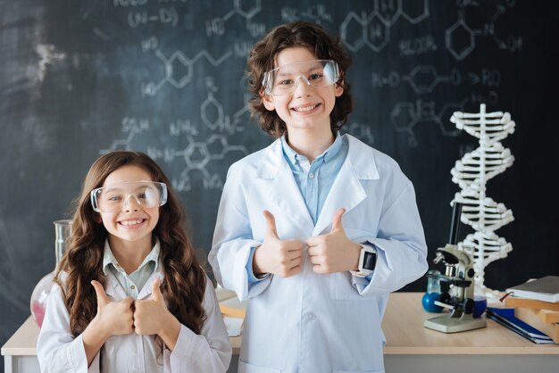 Compartilhando emoções positivas. Sorrindo contentes, pequenos cientistas em pé no laboratório enquanto trabalhavam no projeto e mostrando os polegares para cima