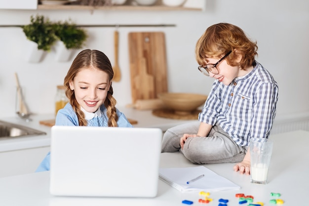 Compartilhando experiência. Garoto atraente encantador e envolvente observando seu irmão estudando para o teste usando seu computador enquanto está sentado ao lado dela na mesa