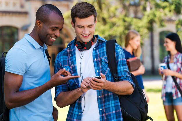 Compartilhando notícias com amigos. Dois homens alegres olhando para o celular e sorrindo com duas mulheres conversando ao fundo