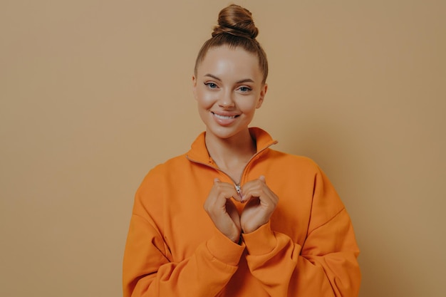 Compartilhe o amor Sorrindo jovem grata fazendo gesto de coração expressando sentimentos calorosos ao namorado isolado no fundo do estúdio bege voluntária feminina mostrando sinal de coração Conceito de caridade