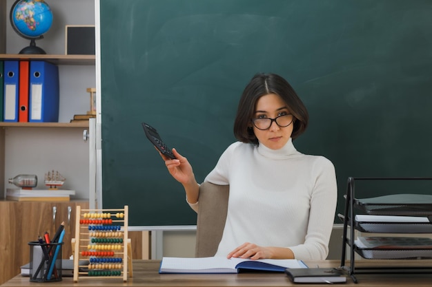 complace mirar a la cámara joven maestra con gafas sosteniendo una calculadora sentada en el escritorio con herramientas escolares en el aula