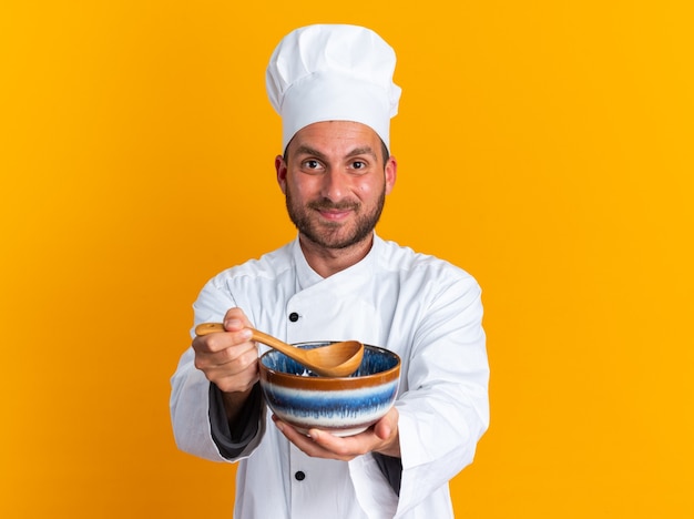 Complacido joven cocinero de sexo masculino caucásico en uniforme de chef y gorra estirando el tazón y la cuchara hacia la cámara