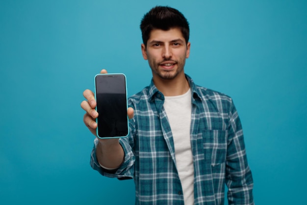 Complacido joven mirando a la cámara estirando el teléfono móvil hacia la cámara aislado sobre fondo azul.