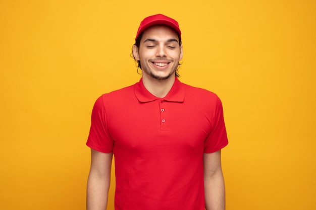 complacido joven repartidor con uniforme y gorra sonriendo con los ojos cerrados aislado sobre fondo amarillo