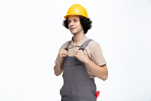 Complacido joven trabajador de la construcción con uniforme y casco de seguridad agarrando su uniforme mirando al lado con instrumentos de construcción en su bolsillo aislado sobre fondo blanco.