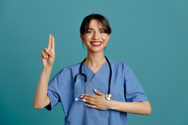 Complacido mostrando gesto joven doctora vistiendo uniforme fith estetoscopio aislado sobre fondo azul.