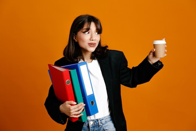 Complacido sosteniendo carpetas con taza de café joven hermosa mujer vistiendo chaqueta negra aislado sobre fondo naranja