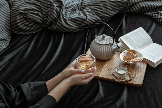 Composição aconchegante com uma xícara de chá nas mãos femininas, biscoitos e um livro na cama