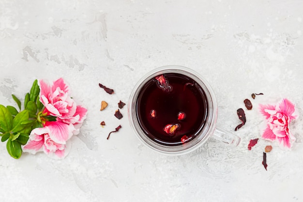 Composição com chá quente de hibisco em caneca de vidro com flores cor de rosa e folhas de chá secas. Vista do topo.