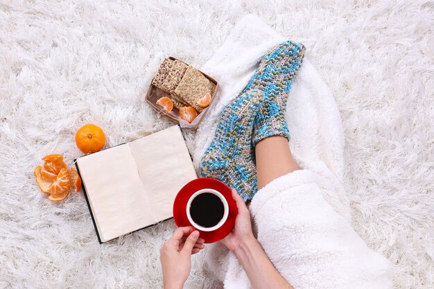 Composição com manta quente, livro, copo de bebida quente e pernas femininas, na cor de fundo do tapete