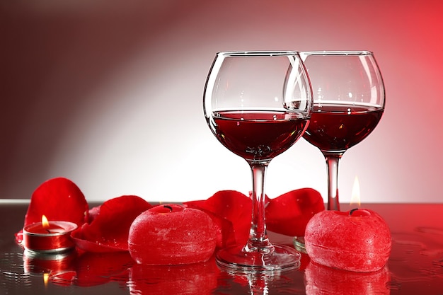 Composição com vinho tinto em copos, pétalas de rosas vermelhas e coração decorativo em fundo colorido