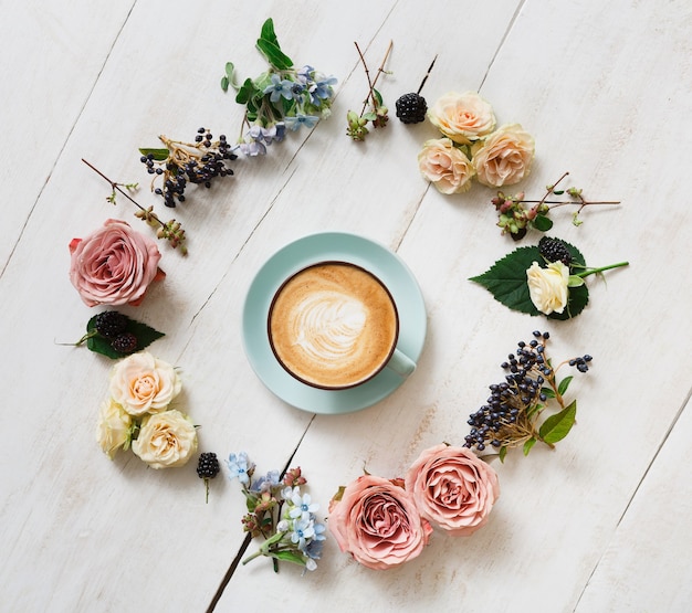 Composição de cappuccino e flores. Xícara de café azul com espuma cremosa, círculo de flores frescas e secas na mesa de madeira branca, vista superior. Bebidas quentes, conceito de oferta sazonal