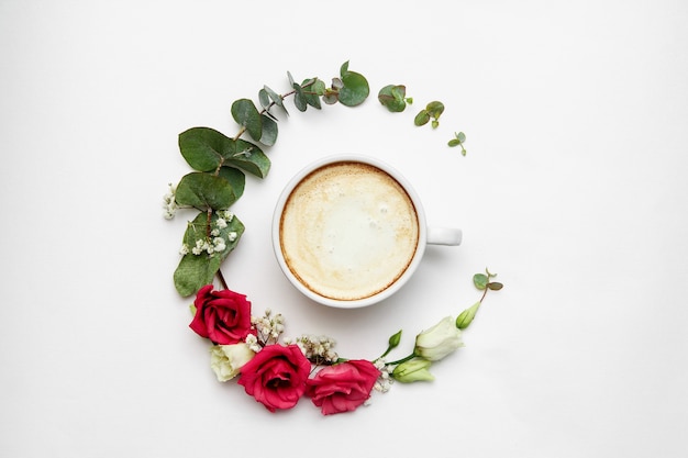 Composição de cappuccino e flores. xícara de café branca com espuma cremosa, círculo de flores frescas em branco, vista superior. bebidas quentes, conceito de oferta sazonal.