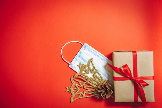 Composição de decorações de Natal, uma caixa de presente em uma fita e uma máscara médica protetora sobre um fundo vermelho.