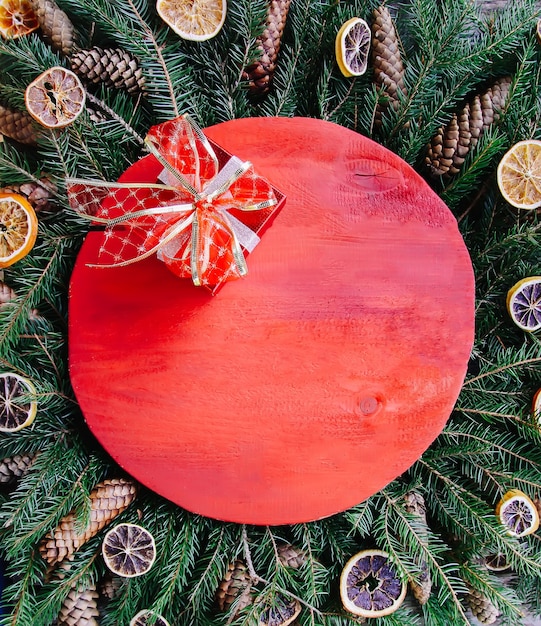 Composição de férias de inverno de Natal e ano novo com superfície de madeira vermelha em galhos de árvore verde do abeto, fatias de laranja secas, cones, caixa de presente e laço vermelho.