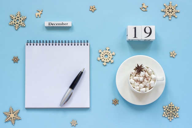 Composição de inverno. Calendário de madeira 19 de dezembro Xícara de cacau com marshmallow e anis estrelado, bloco de notas aberto vazio no conceito Mockup leigo