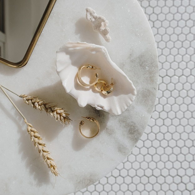 Composição de moda mínima com brincos de ouro em concha do mar na mesa de mármore com espelho e hastes de trigo.