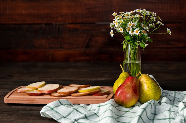 Composição de natureza morta com vaso de flores silvestres, toalha de linho de cozinha, peras maduras e tábua de cortar com fatias de frutas frescas