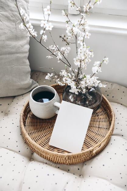 Composição de natureza morta de primavera Maquete de cartão na bandeja de vime xícara de café Foto com estilo feminino Cena floral com flores de cerejeira branca borrada no banco perto da janela Foco seletivo