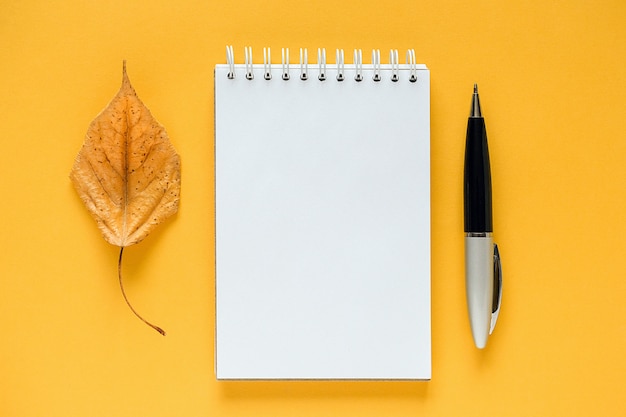 Composição de outono. Bloco de notas em branco branco, folha alaranjada secada e pena no amarelo.