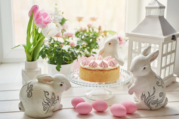 Composição de páscoa com bolo doce com glacê de morango, coelhinhos de cerâmica, ovos cor de rosa e rosas