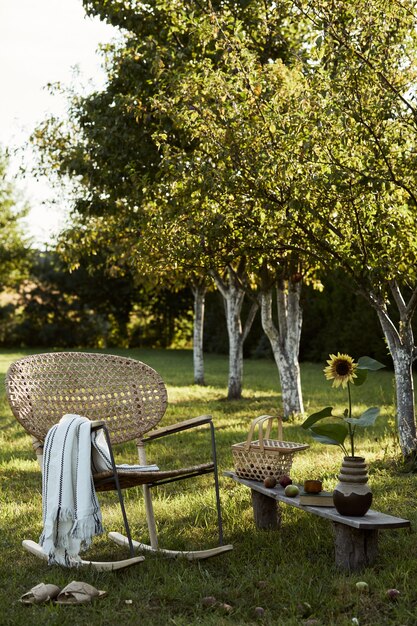 Composição elegante de jardim rural com poltrona de vime de design, banco de madeira, xadrez, alimentos, bebidas e acessórios elegantes. Muitas flores coloridas. Clima de verão.