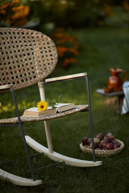 Composição elegante de jardim rural com poltrona de vime de design, banco de madeira, xadrez, alimentos, bebidas e acessórios elegantes. muitas flores coloridas. clima de verão.