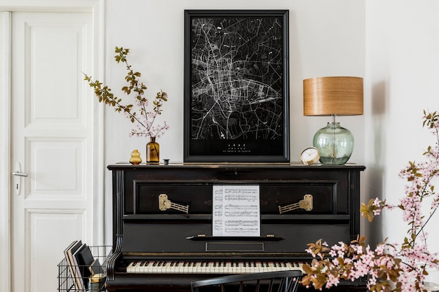 Composição elegante no interior da sala de estar com piano preto, mapa de pôster, flores secas, relógio de ouro, lâmpada de design e acessórios pessoais elegantes em decoração moderna.