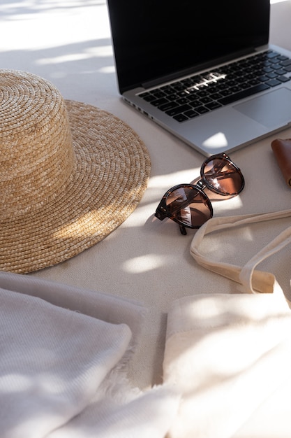 Composição estética criativa com acessórios de moda feminina. óculos de sol femininos elegantes, chapéu de palha, bolsa de compras, laptop no sofá branco com almofadas