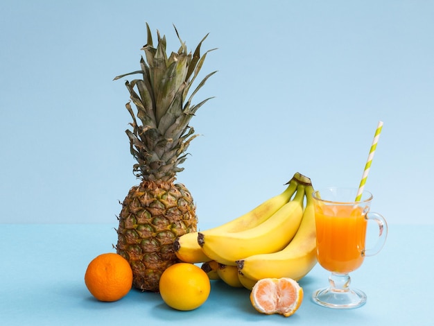 Composição natural de frutas tropicais. Abacaxi fresco, banana, tangerina e limão com um copo de suco de fruta sobre fundo azul.