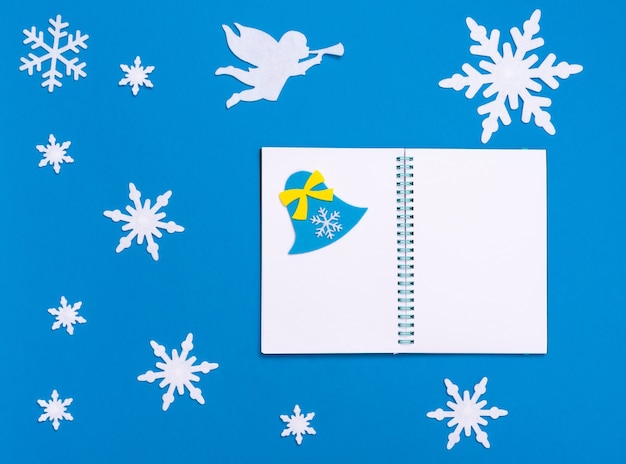 Composição plana de natal e ano novo com bloco de notas em branco branco, sino azul, anjo branco tocando trompete e flocos de neve de natal em fundo azul