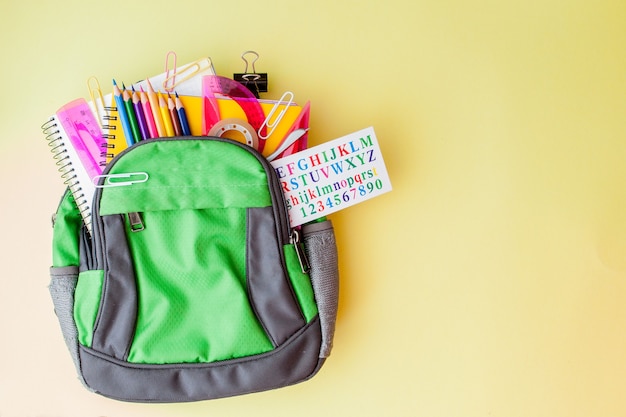 Foto composição plana leiga com mochila e papelaria escolar em fundo amarelo.