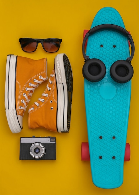 Composição plana leiga de acessórios de jovens hipster. placa cruzadora, tênis, fones de ouvido, câmera e óculos de sol em fundo amarelo. vista do topo