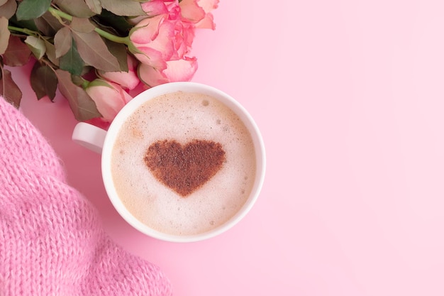 Composição romântica com xícara de café com coração em cima de flores e cobertor de malha em fundo rosa pastel