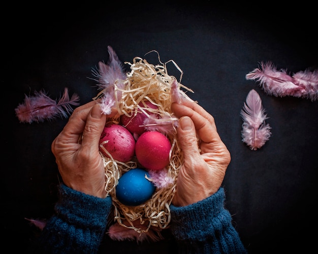 Composición alternativa de Pascua en estilo oscuro con huevos de color azul y rosa clásico, plumas y manos de mujer mayor sobre superficie negra, concepto de vacaciones de temporada de primavera