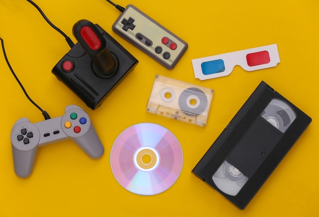 Composición de atributos plana de los años 80. Gamepads, cintas de audio y video, CD, gafas 3D. Electrónica retro, gadgets y dispositivos. Entretenimiento y juegos. Fondo amarillo. Vista superior