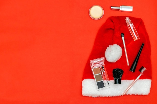 Composición de cosméticos de belleza de año nuevo con pinceles, lápiz labial, sombra de ojos, polvo, accesorios, decoraciones de bolas sobre fondo rojo de sombrero de navidad. Vista superior de maqueta plana elegante con espacio de copia