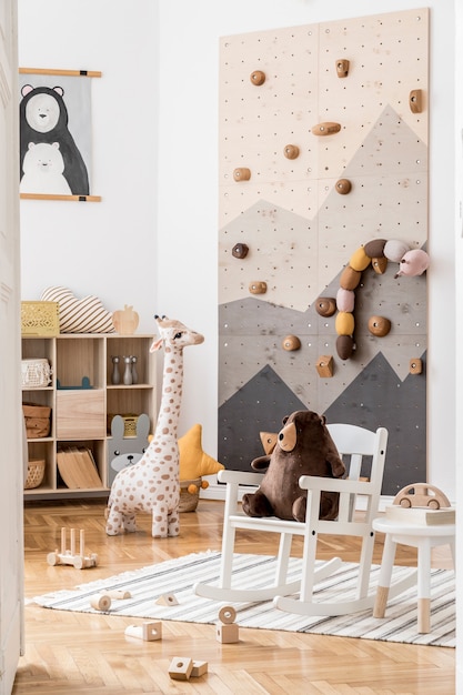 Foto composición creativa del interior de la habitación infantil scandi elegante y acogedor con gráfico en la pared, juguetes y juguetes de peluche, sofá, inodoro, sillón, alfombra y accesorios paredes blancas y piso de parquet