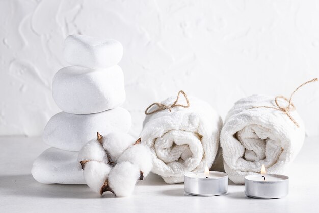 Composición creativa con piedras zen, toallas enrolladas, velas y flores de algodón sobre fondo blanco.