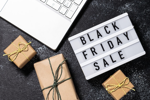 Composición creativa con texto de Black Friday Sale en la caja de luz, el portátil y las cajas de regalo en papel artesanal sobre fondo rústico negro. Concepto de compra online.